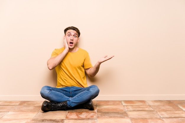 Young caucasian man sitting on the floor isolated holds copy space on a palm, keep hand over cheek. Amazed and delighted.