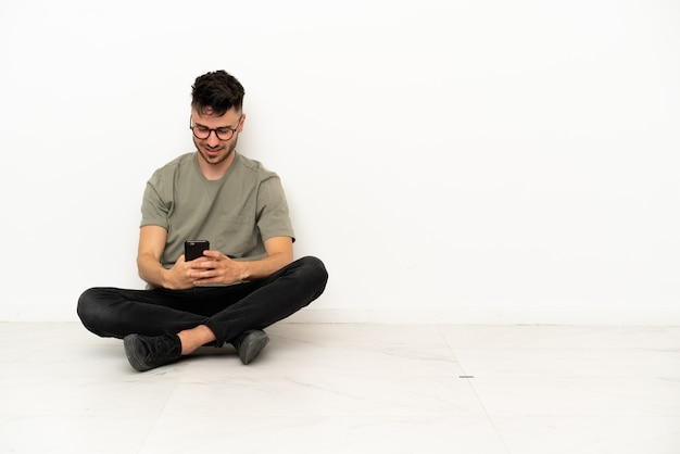 Young caucasian man sitting on the floor isolated on white background sending a message with the mobile