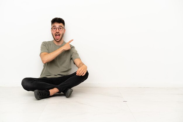 Young caucasian man sitting on the floor isolated on white background surprised and pointing side