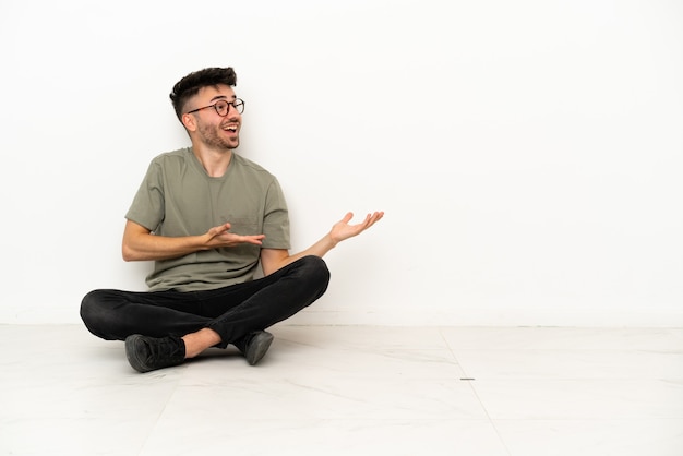 Молодой кавказский мужчина сидит на полу на белом фоне с удивленным выражением лица
