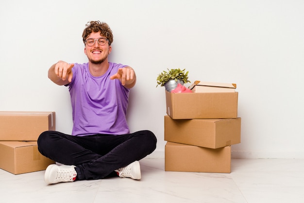 Young caucasian man sitting on the floor ready for moving isolated on white background cheerful smiles pointing to front.