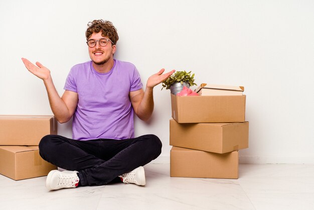 Young caucasian man sitting on the floor ready for moving isolated on white background doubting and shrugging shoulders in questioning gesture.