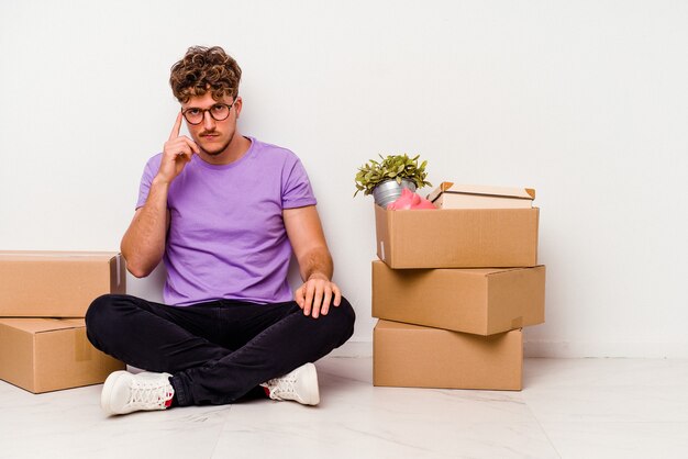 Young caucasian man sitting on the floor ready for moving isolated on white background pointing temple with finger, thinking, focused on a task.