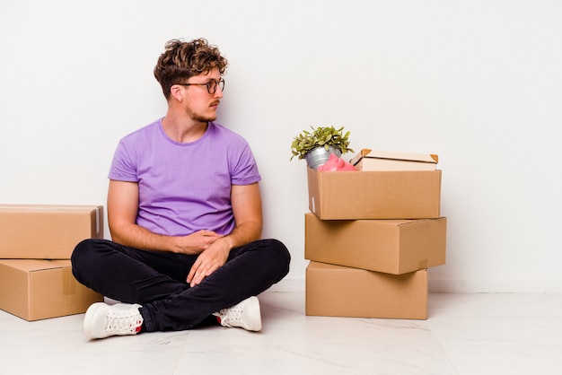 Young caucasian man sitting on the floor ready for moving isolated on white wall gazing left, sideways pose.