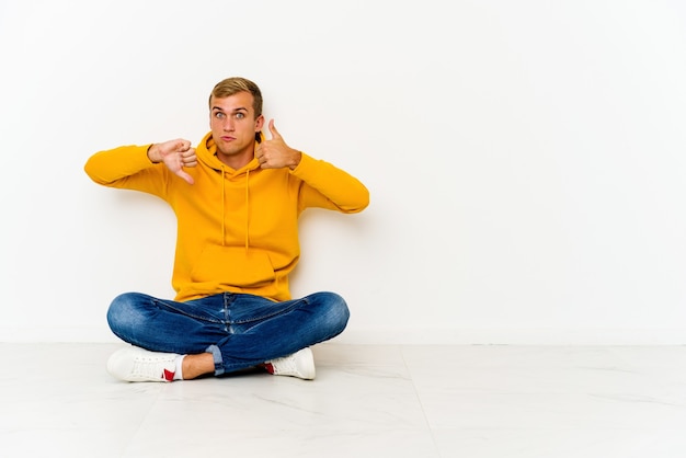 Young caucasian man sitting on the floor showing thumbs up and thumbs down, difficult choose concept