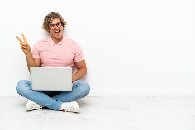 Young caucasian man sitting on the floor with his laptop isolated on white wall smiling and showing victory sign