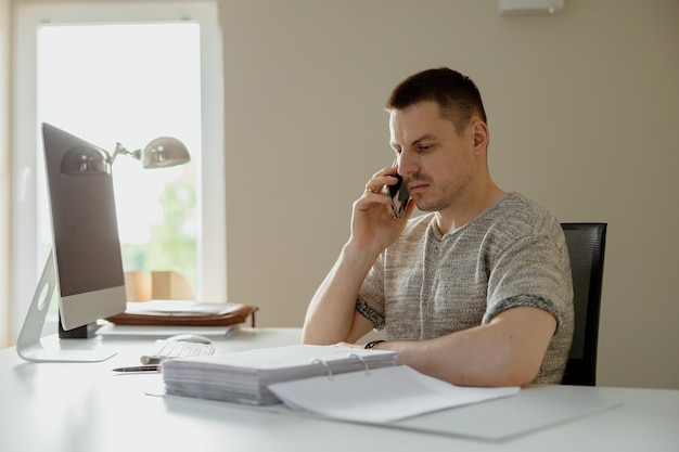 Young caucasian man sitting at his desk in office working with computer and communicating over smartphone Young employee has a working day in office Entrepreneur talks on a phone about his business