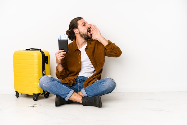Молодой кавказский человек сидит с паспортом и чемоданом, кричит и держит ладонь возле открытого рта.