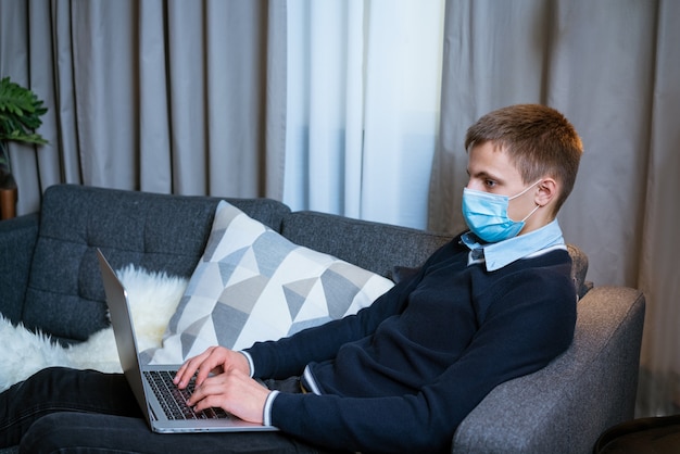 Young caucasian man sitting at home on sofa with laptop in medical mask