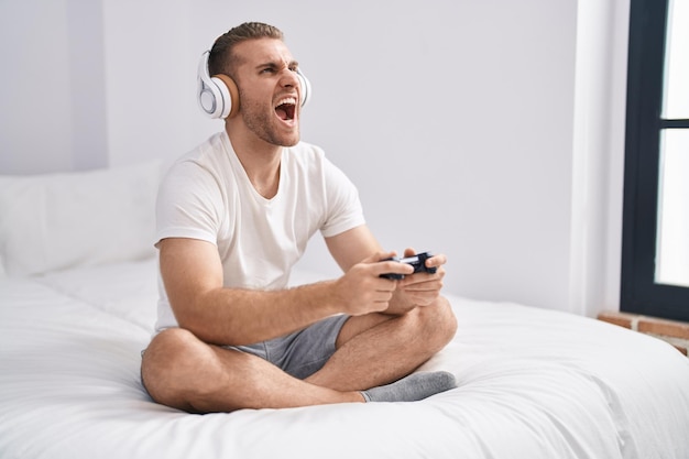 Фото Молодой белый мужчина сидит на кровати дома, играет в видеоигры, злится и злится, кричит, расстроен и яростен, кричит от гнева, смотрит вверх.