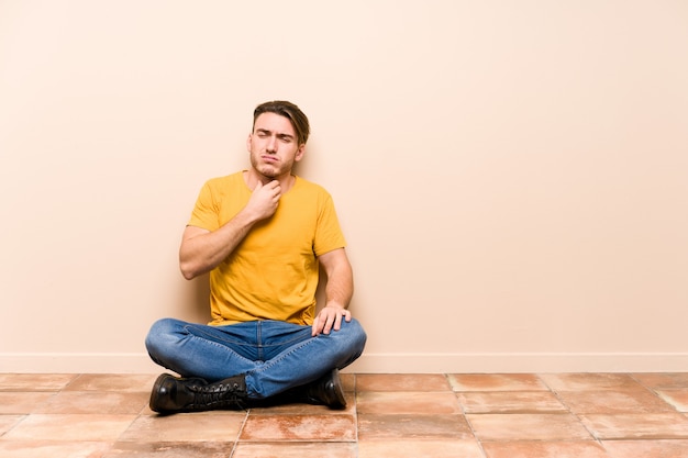 사진 고립 된 바닥에 앉아 젊은 백인 남자는 바이러스 또는 감염으로 인해 목에 통증을 앓고 있습니다.
