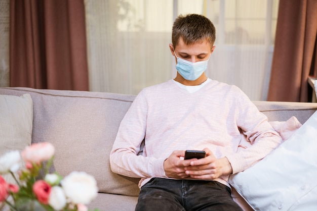 Young caucasian man sitting on a sofa in casual clothes wearing a protective medical mask with a pho...