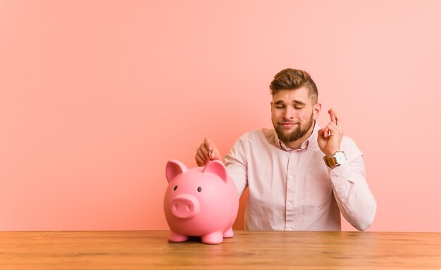 Young caucasian man sitting with a piggy bank crossing fingers for having luck