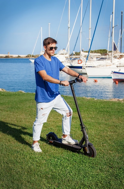 Il giovane uomo caucasico rimane con uno scooter elettrico sull'erba vicino al mare nel porto turistico