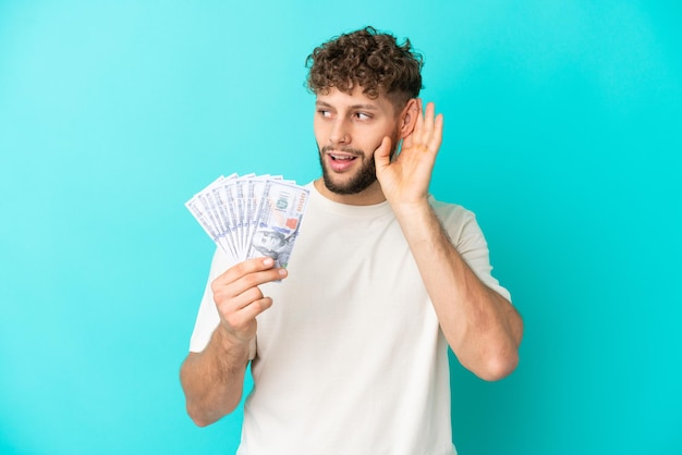Young caucasian man taking a lot of money isolated on blue background listening to something by putting hand on the ear