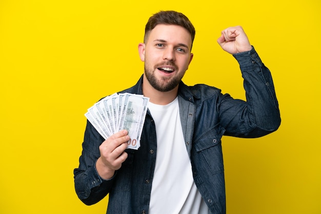 Young caucasian man taking a lot of money isolated on yellow background celebrating a victory