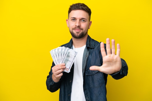 Young caucasian man taking a lot of money isolated on yellow background counting five with fingers