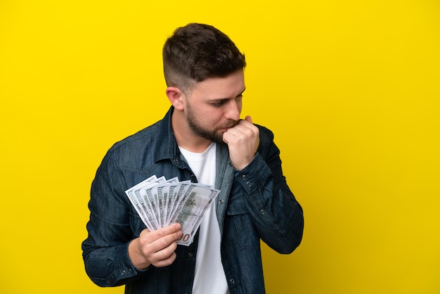 Young caucasian man taking a lot of money isolated on yellow background having doubts