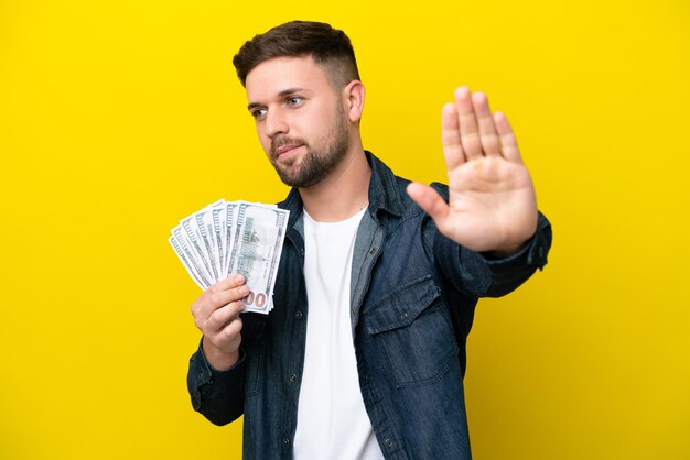 Young caucasian man taking a lot of money isolated on yellow background making stop gesture and disappointed