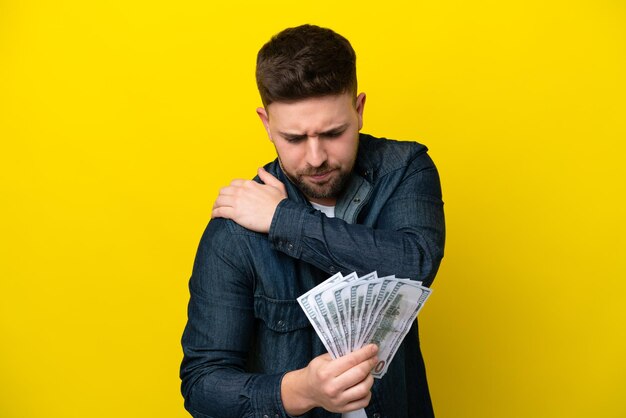 Young caucasian man taking a lot of money isolated on yellow background suffering from pain in shoulder for having made an effort