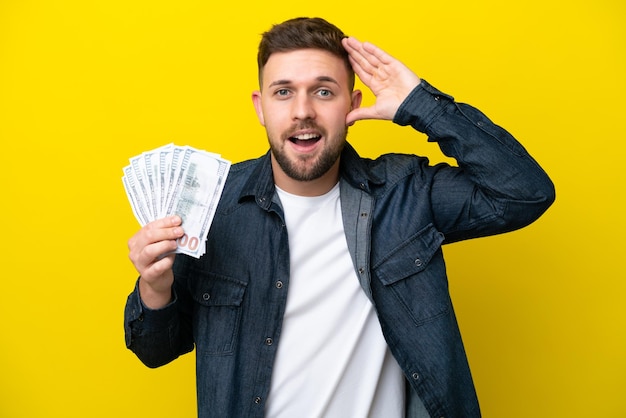 Young caucasian man taking a lot of money isolated on yellow background with surprise expression