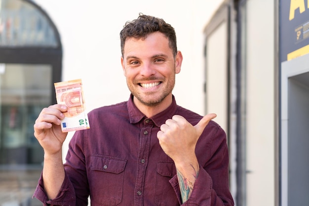 young caucasian man taking a lot of money at outdoors pointing to the side to present a product