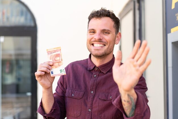 Young caucasian man taking a lot of money at outdoors saluting with hand with happy expression