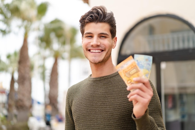 Young caucasian man taking a lot of money at outdoors smiling a lot