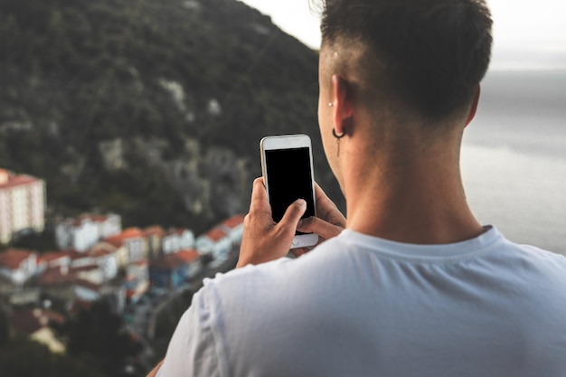 Giovane uomo caucasico che scatta una foto con un telefono cellulare da una vista dall'alto di una piccola città costiera