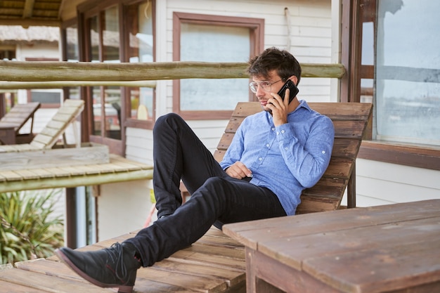 Young Caucasian man talking on his smart phone while resting on a wooden bench on a beautiful terrace of his cabin.