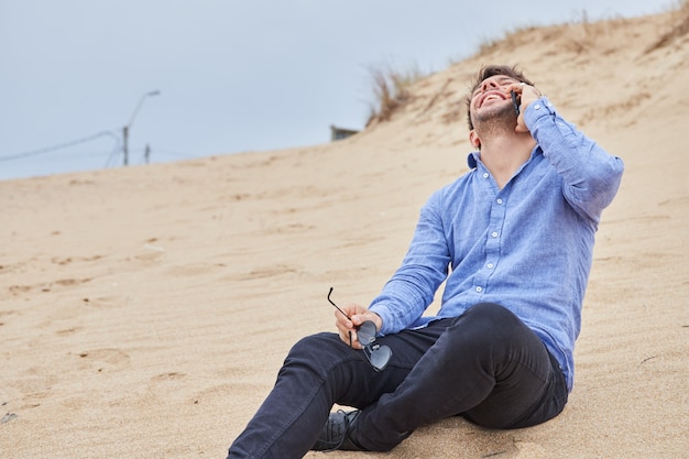 Young Caucasian man talking on the phone and smiling sitting on the sand on the beach. He is wearing a light blue shirt, black pants and black shoes.