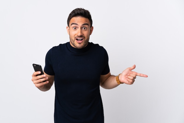 Young caucasian man using mobile phone isolated on white wall surprised and pointing finger to the side