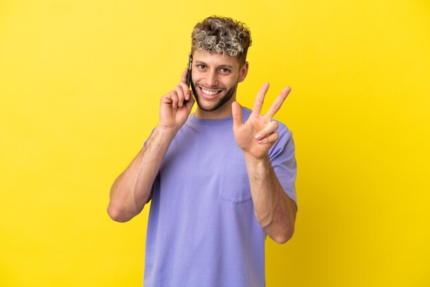 Young caucasian man using mobile phone isolated on yellow background happy and counting three with fingers