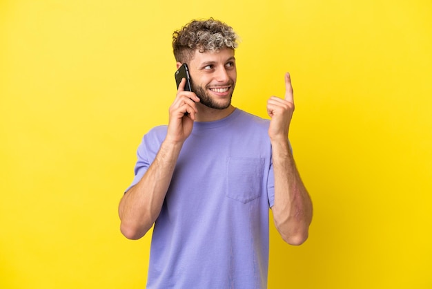 Young caucasian man using mobile phone isolated on yellow background pointing up a great idea