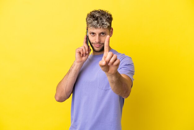 Young caucasian man using mobile phone isolated on yellow background showing and lifting a finger