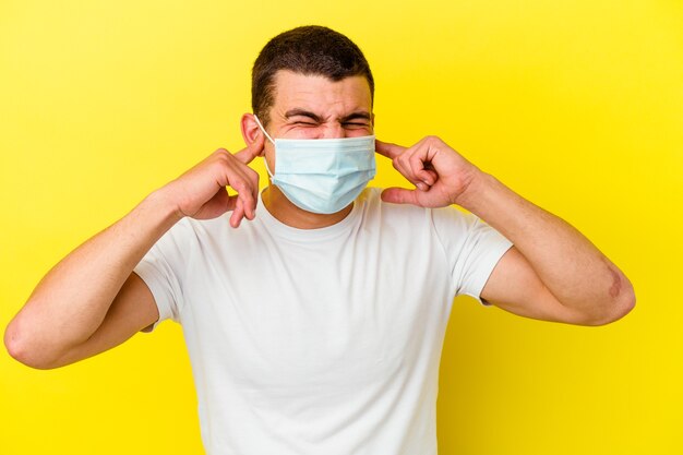 手で耳を覆う黄色の背景に分離されたコロナウイルスの保護を身に着けている若い白人男性。