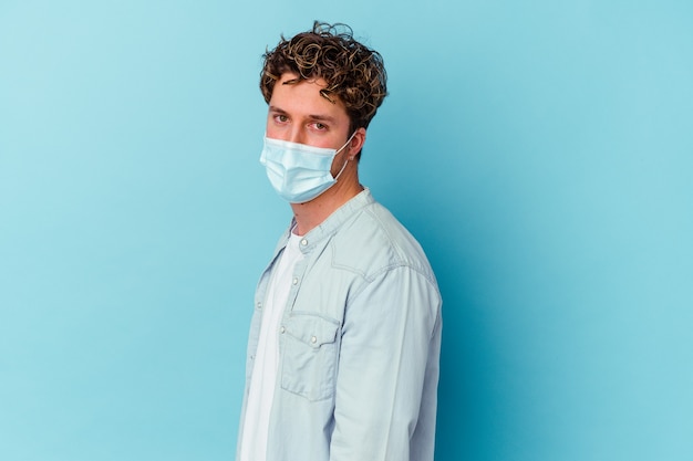 Young caucasian man wearing an antiviral mask isolated on blue background looks aside smiling, cheerful and pleasant