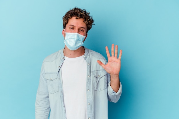 Young caucasian man wearing an antiviral mask isolated on blue background smiling cheerful showing number five with fingers.