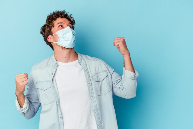 Young caucasian man wearing an antiviral mask isolated on blue wall raising fist after a victory, winner concept.