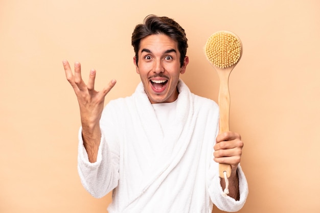 Photo young caucasian man wearing a bathrobe holding a back scratcher isolated on beige background receiving a pleasant surprise, excited and raising hands.