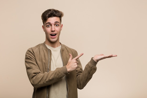 Young caucasian man wearing a brown jacket excited holding a copy space on palm.