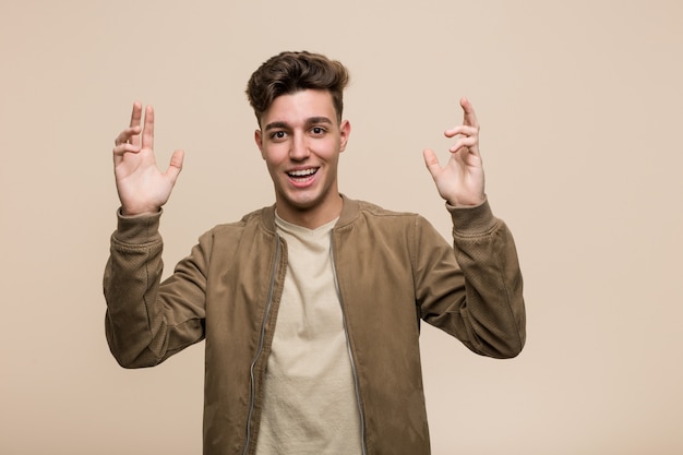 Young caucasian man wearing a brown jacket receiving a pleasant surprise, excited and raising hands.