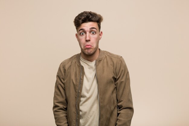Young caucasian man wearing a brown jacket shrugs shoulders and open eyes confused.