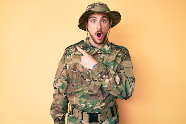 Young caucasian man wearing camouflage army uniform surprised pointing with finger to the side open mouth amazed expression