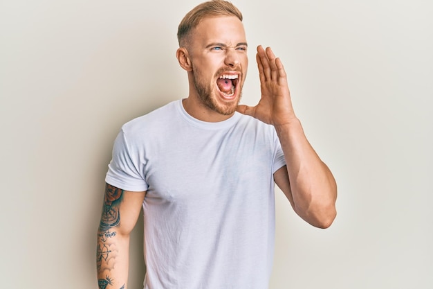 Young caucasian man wearing casual white tshirt shouting and screaming loud to side with hand on mouth communication concept