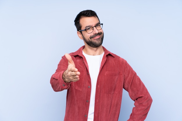 Young caucasian man wearing corduroy jacket over blue wall shaking hands for closing a good deal