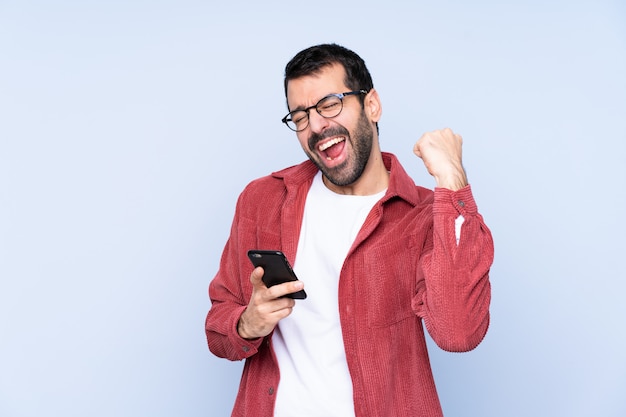 Young caucasian man wearing corduroy jacket over blue wallwith phone in victory position