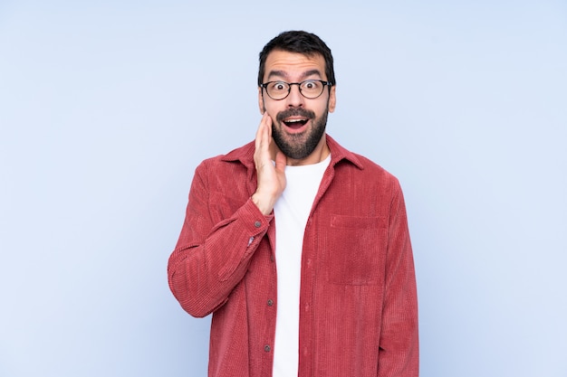 Young caucasian man wearing corduroy jacket over blue wallwith surprise and shocked facial expression