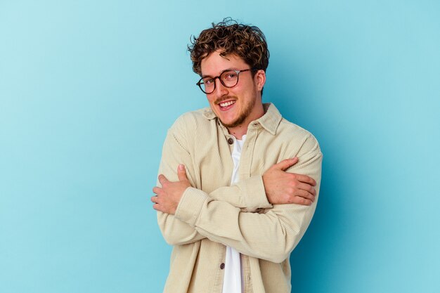 Young caucasian man wearing eyeglasses isolated on blue background hugs, smiling carefree and happy.