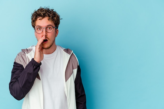 Young caucasian man wearing eyeglasses isolated on blue background is saying a secret hot braking news and looking aside